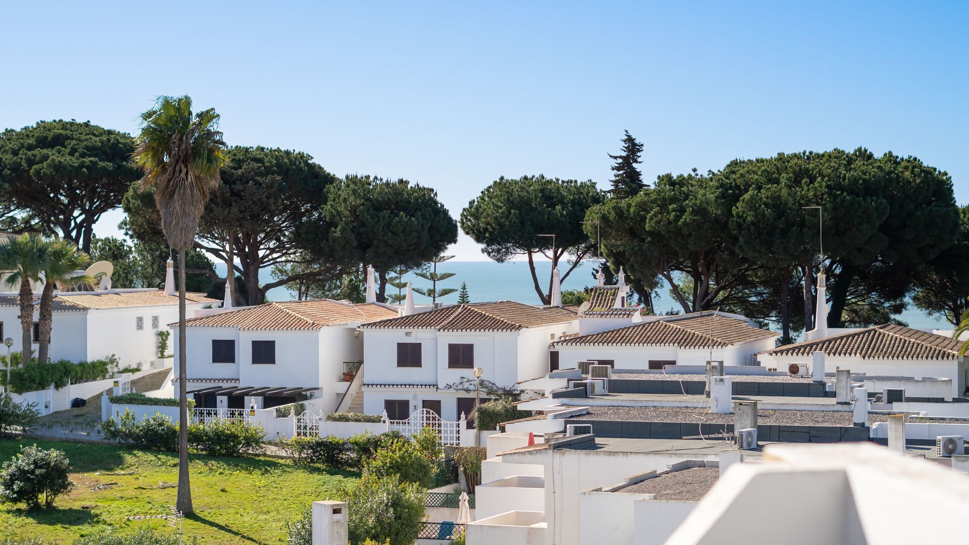 A coastal residential area with white buildings, some with tiled roofs, surrounded by greenery and trees.