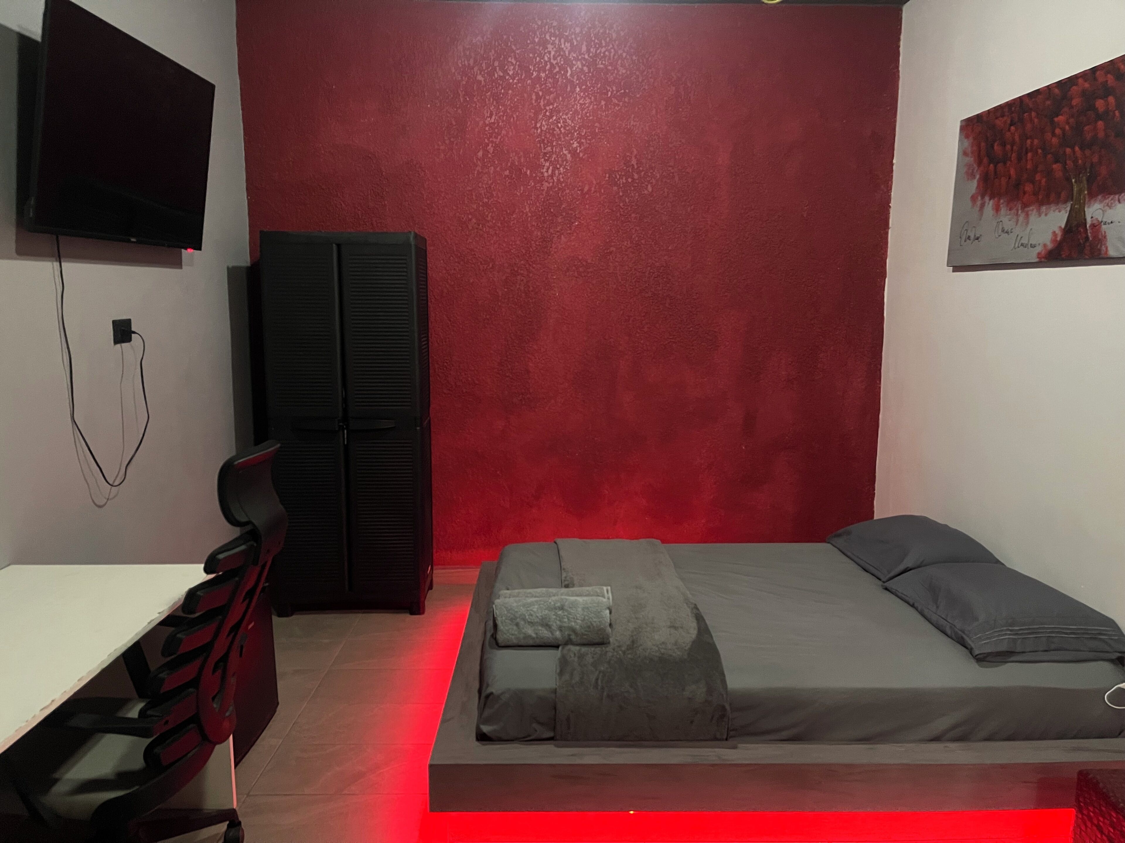 Een slaapkamer met een rode accentmuur, een bed met grijze beddengoed, een bureau met een stoel, een zwarte garderobe en een aan de muur bevestigde televisie.