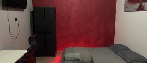 Een slaapkamer met een rode accentmuur, een bed met grijze beddengoed, een bureau met een stoel, een zwarte garderobe en een aan de muur bevestigde televisie.
