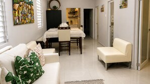 Un salon moderne comprenant un coin repas, un canapé, une table basse et une table à manger avec des chaises. Un tableau orne le mur, un luminaire est suspendu au plafond et une porte donne accès à une autre pièce.