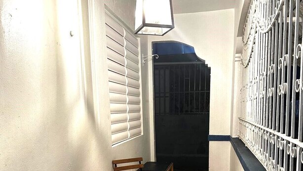 Un pasillo estrecho con suelo de baldosas, una mesa y sillas de madera, una lámpara colgante y una verja.