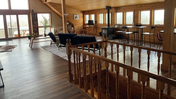 Um amplo quarto estilo loft, com teto alto, vigas de madeira e grandes janelas. Há uma área de estar aconchegante com um sofá e cadeiras, uma mesa de jantar e uma lareira.