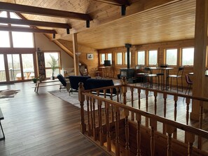 Amplia habitación estilo loft con techo alto, vigas de madera y ventanas grandes. Cuenta con una zona de estar acogedora con sofá y sillones, una mesa de comedor y una chimenea.