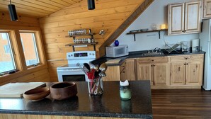 Cozinha com armários de madeira, fogão, micro-ondas e cafeteira. Há duas janelas e teto de madeira.