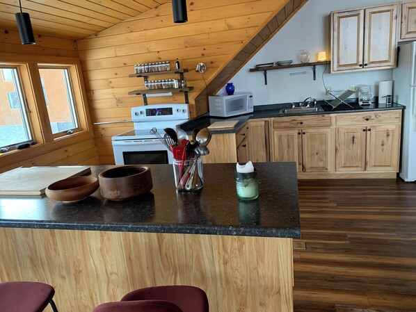 Una cocina con armarios de madera, una estufa, un microondas y una cafetera. Hay dos ventanas y un techo de madera.