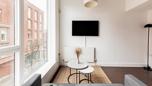 Uma sala de estar moderna com uma janela grande, uma TV de tela plana na parede, uma pequena mesa redonda com um vaso e um abajur.