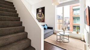 A staircase leading to a living area with a sofa, a small round table, and a large window offering a view of buildings. - Stylish Camden Flat Near Markets & Tube (London)