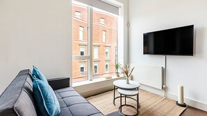 A modern living room with a grey sofa, a small round table, and a flat-screen TV mounted on the wall. - Stylish Camden Flat Near Markets & Tube (London)
