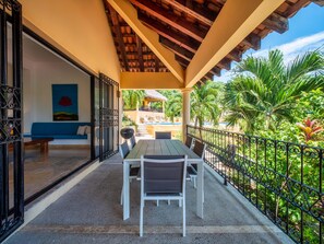 A covered patio with a table and chairs, a blue sofa, and a view of a pool and greenery.