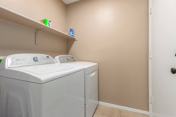 A laundry room with a front-loading washer and dryer, a shelf above the dryer with cleaning supplies, and a beige wall. - Charming 3-bedroom house with WiFi, AC, Private Pool in pleasant Surprise (Surprise)