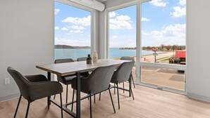 Ein Essbereich mit großem Tisch und Stühlen, große Fenster mit Blick auf ein Gewässer und eine Straße mit Fahrzeugen.