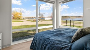 A bedroom with a large window offering a view of a road, trees, and a body of water.
