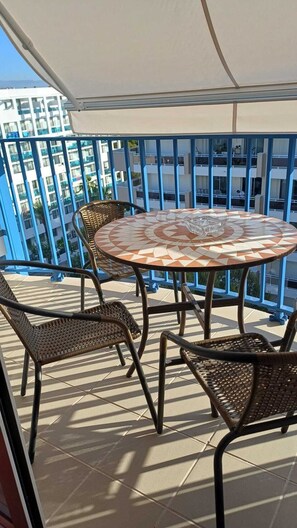 Une table ronde recouverte d’une nappe à motifs, entourée de chaises en osier, sur un balcon doté d’une rambarde bleue et offrant une vue sur un immeuble.