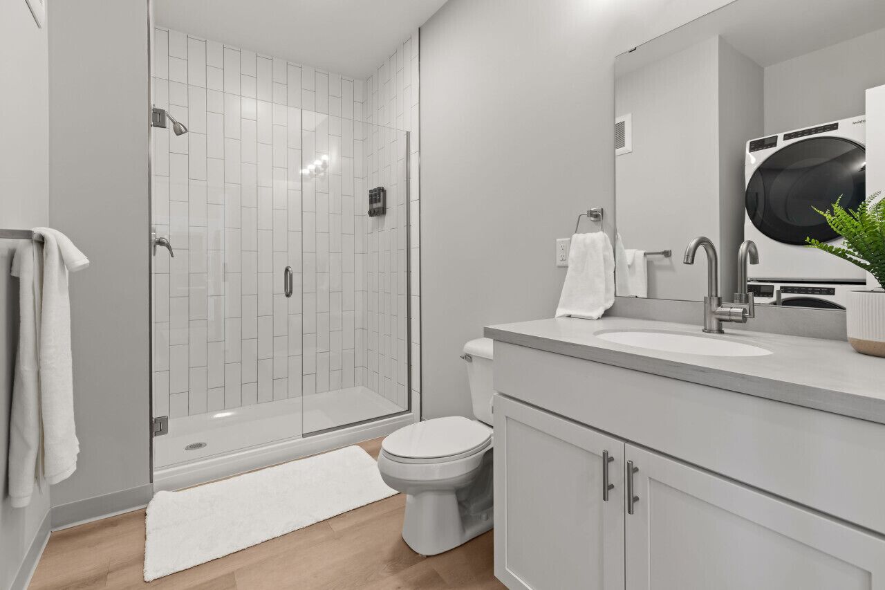 Een moderne badkamer met een glazen douchecabine, een toilet, een wastafel met grote spiegel en een wandmodel wasmachine.