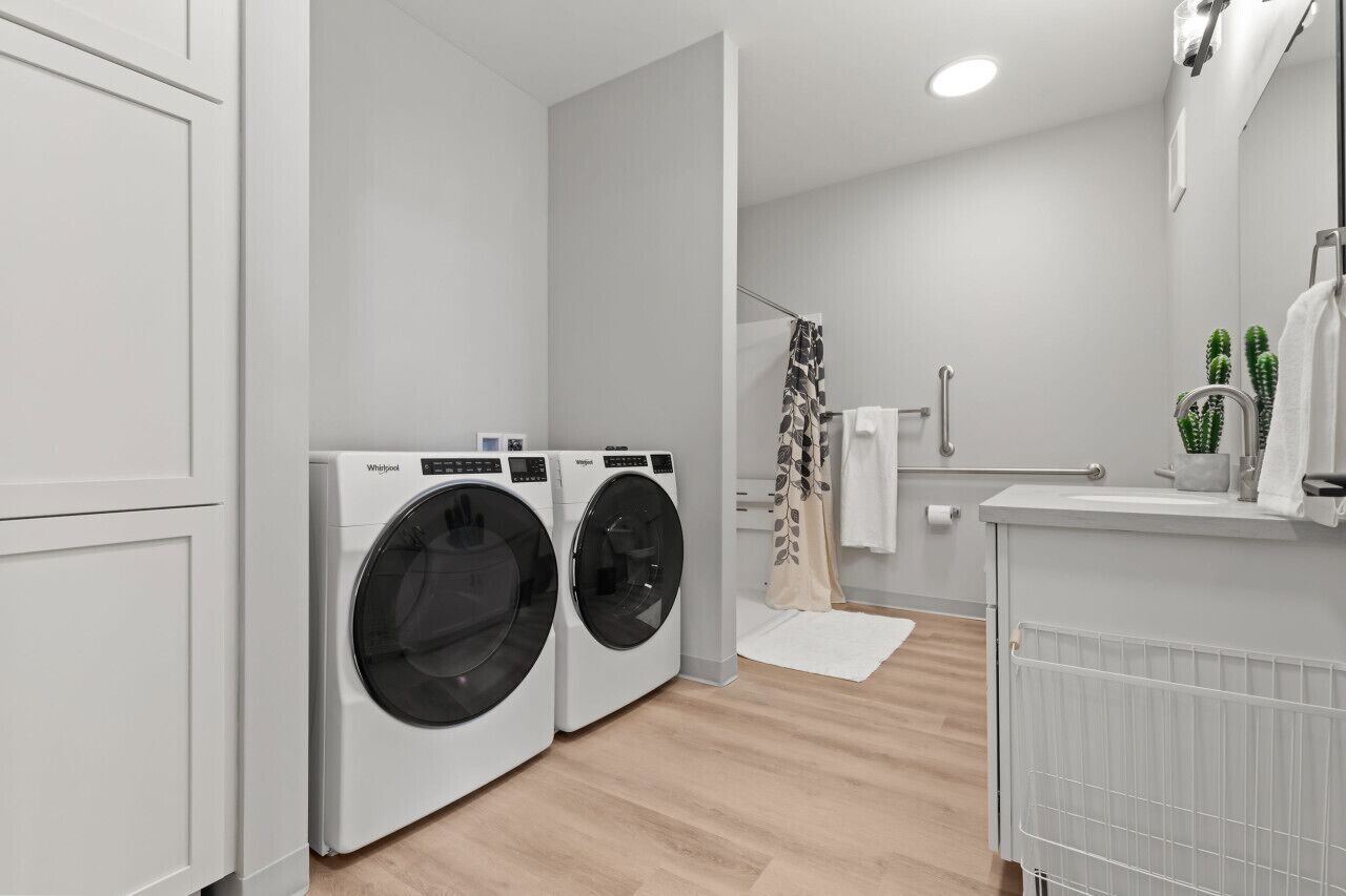 A modern laundry room with two front-loading washers and dryers, a shower area with a glass door, and a white sink with a mirror above it.