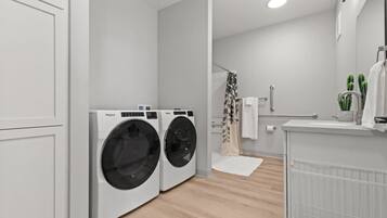 A modern laundry room with two front-loading washers and dryers, a shower area with a glass door, and a white sink with a mirror above it.