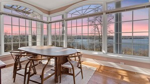 Uma sala de jantar com uma mesa grande e cadeiras, janelas amplas com vista para um lago e árvores, e piso de madeira.