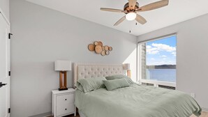 Un dormitorio con cama, mesita de noche, ventilador de techo, ventana con vista al agua y decoración en la pared.