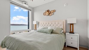 A bedroom with a large window, a tufted headboard, two bedside tables with lamps, and a wall-mounted decorative piece.