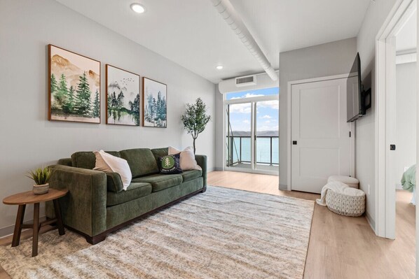 Een moderne woonkamer met een groene bank, een klein rond tafeltje met een plant en een groot raam met uitzicht op de omgeving.
