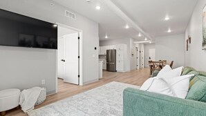 A spacious living area with a green sofa, white cushions, and a white rug. There is a kitchen in the background with white cabinets and a refrigerator.