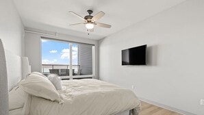 Un dormitorio con cama, ventilador de techo, televisor montado en la pared y una ventana grande con vista al cielo.