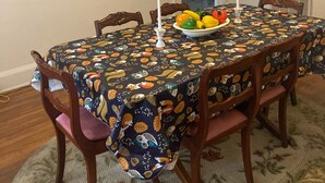 A dining room with a table covered in a tablecloth with a fruit pattern, surrounded by wooden chairs. There is a bowl of fruit on the table, and a candle in the center. The room has a 'home' sign on the wall and a door leading to another room. - Charming 2-bedroom townhome in quaint Hamlet with WiFi, AC (Hamlet)