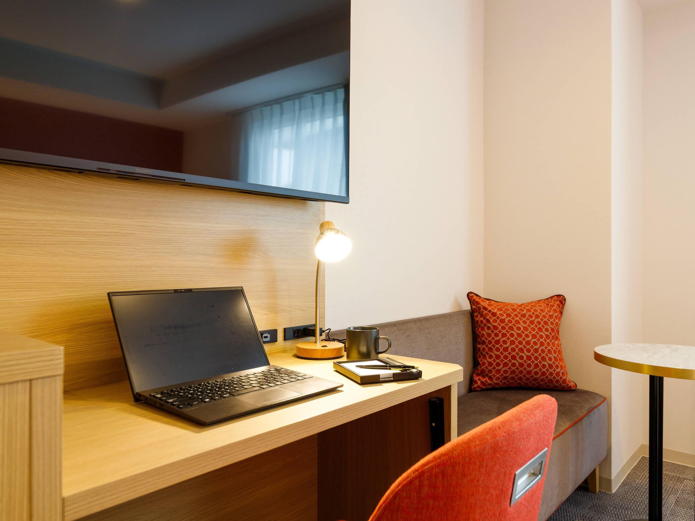 A hotel room with a wooden desk, a laptop, a desk lamp, a small table, a sofa, and a TV mounted on the wall.