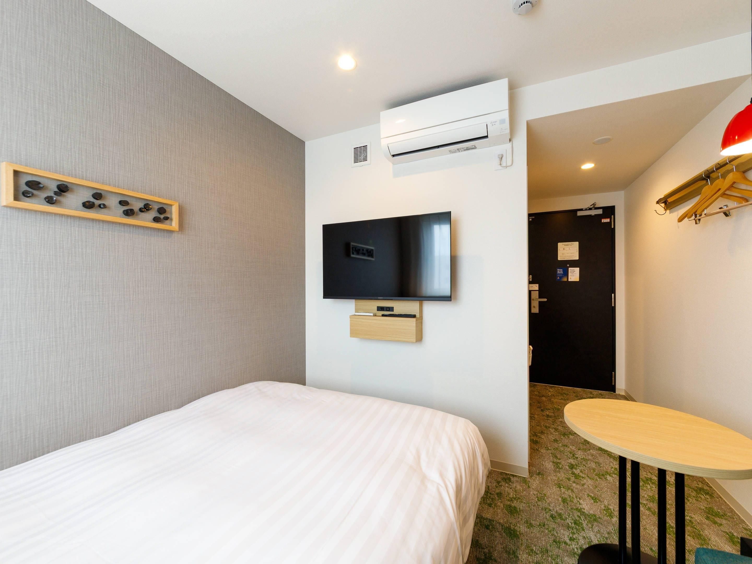 A hotel room with a bed, a mounted TV, an air conditioning unit, a small table, and a wall-mounted shelf with decorative items.