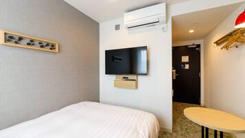 A hotel room with a bed, a mounted TV, an air conditioning unit, a small table, and a wall-mounted shelf with decorative items.