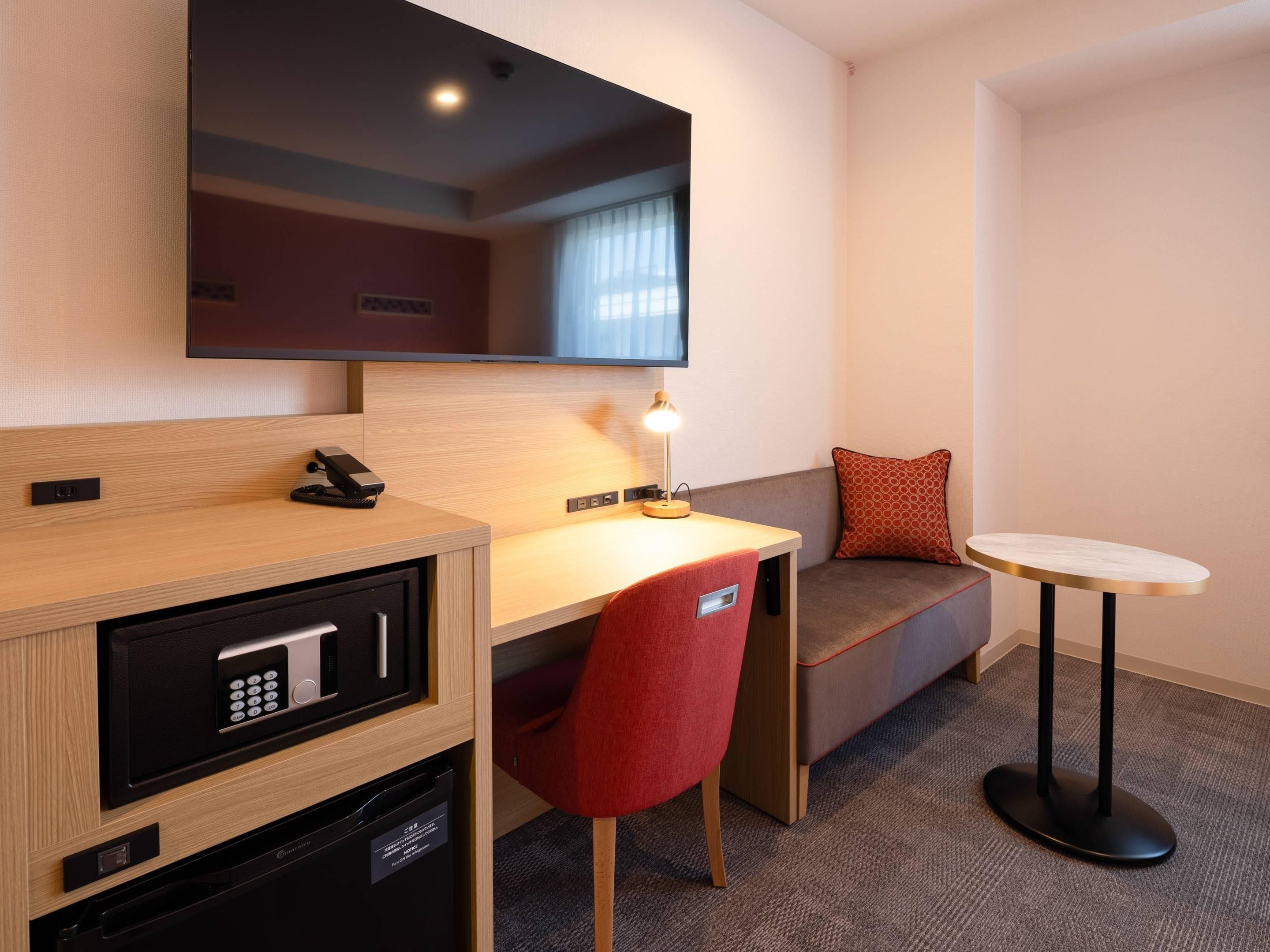 Une chambre d’hôtel compacte, dotée d’un bureau en bois, d’une chaise rouge, d’une petite table ronde, d’un canapé et d’un téléviseur fixé au mur.