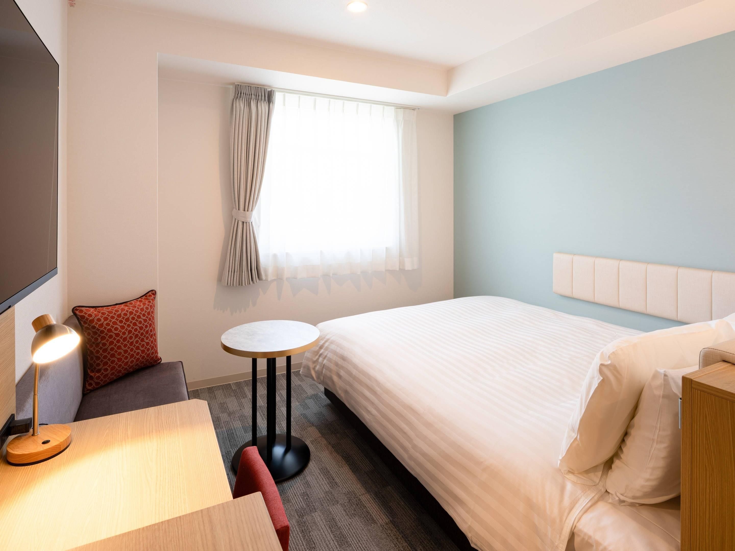 Une chambre d’hôtel avec un lit, une petite table ronde, une lampe, un canapé et une fenêtre avec des rideaux.