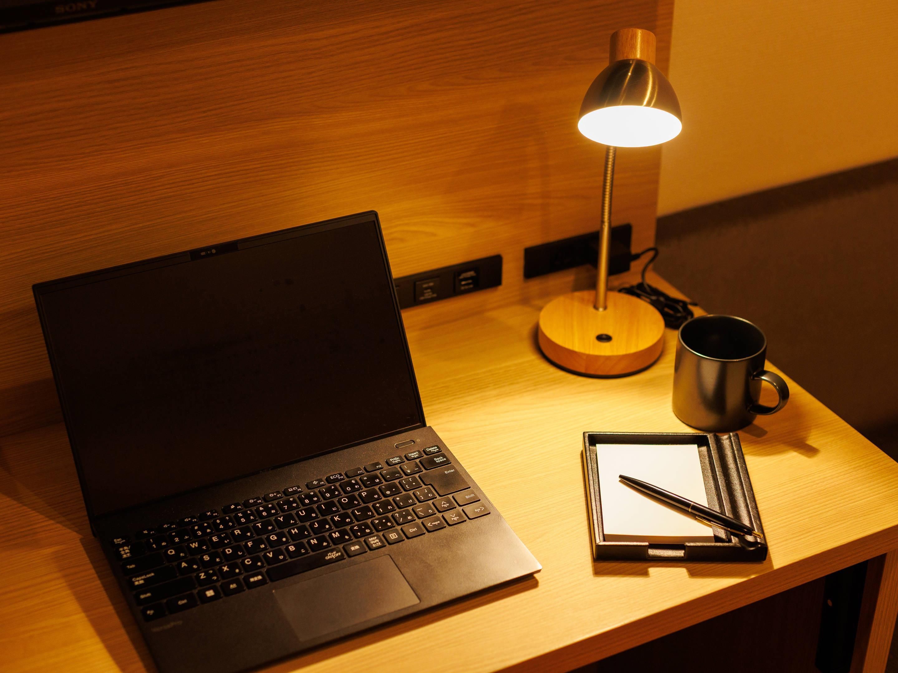 Una computadora portátil sobre un escritorio de madera con una lámpara, una taza y un bloc de notas.