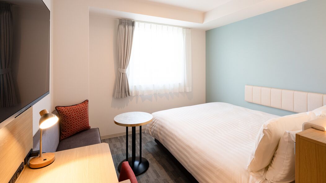 Ein Hotelzimmer mit einem Bett, einem Schreibtisch mit Lampe, einem kleinen runden Tisch, einem Fernseher und einem Fenster mit Vorhängen.