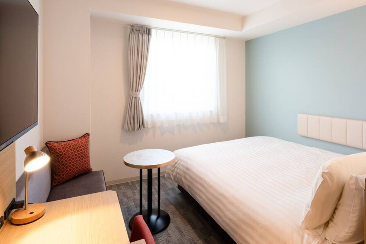 Habitación de hotel con cama, escritorio con lámpara, una mesita redonda, televisor y ventana con cortinas.