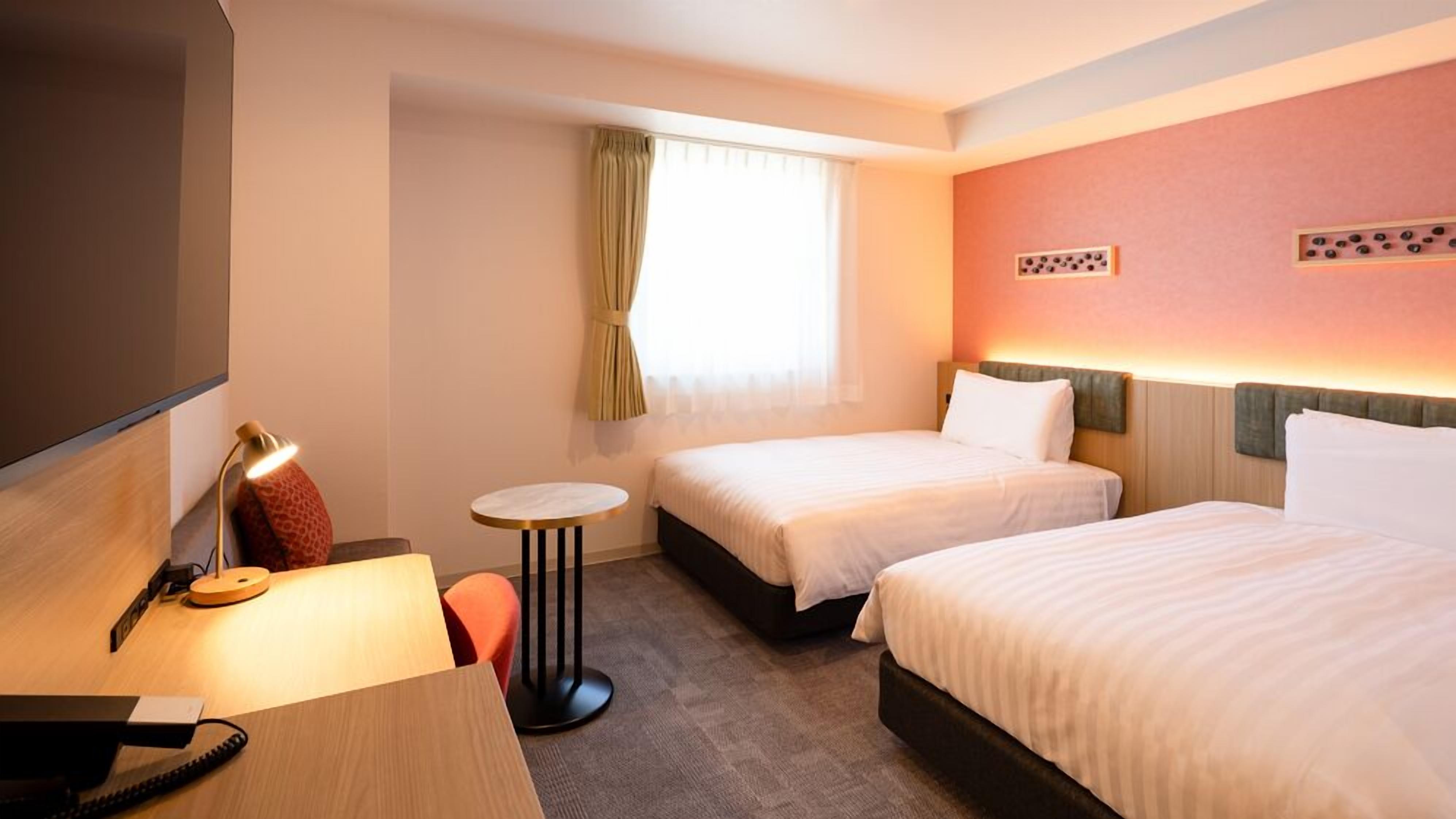 Ein Hotelzimmer mit zwei Betten, einem Schreibtisch mit Lampe, einem kleinen Tisch und einem Fernseher.