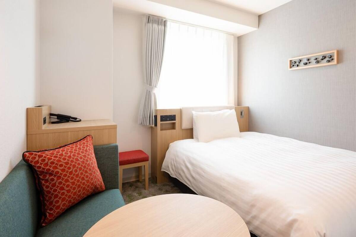 Una habitación de hotel con cama, escritorio, silla, una mesita y ventana con cortinas.