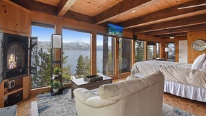 A cozy cabin room with a fireplace, a bed, a sofa, and large windows offering a view of a lake and mountains. - Stunning Views at Stargazer | Lakefront Studio! (Crystal Bay)