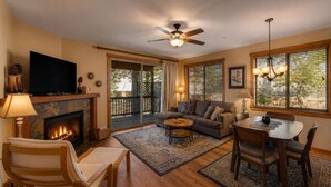 A living room with a fireplace, a television, a ceiling fan, a dining table with chairs, and a large window with blinds.