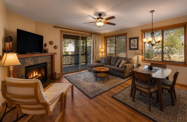A living room with a fireplace, a television, a ceiling fan, a dining table with chairs, and a large window with blinds.