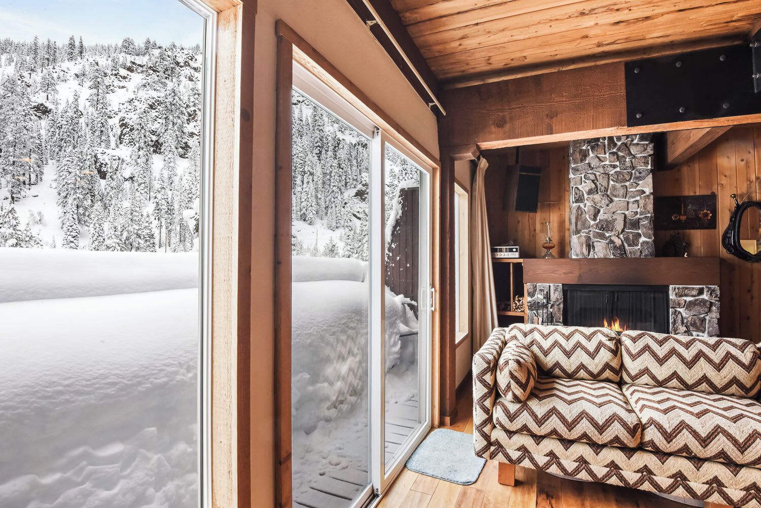 A cozy cabin interior with a stone fireplace, a sofa with patterned cushions, and a view of a snowy forest through large sliding glass doors.