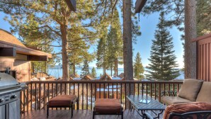 Terraza de madera con mobiliario exterior, una parrilla y vistas a un lago y bosque.