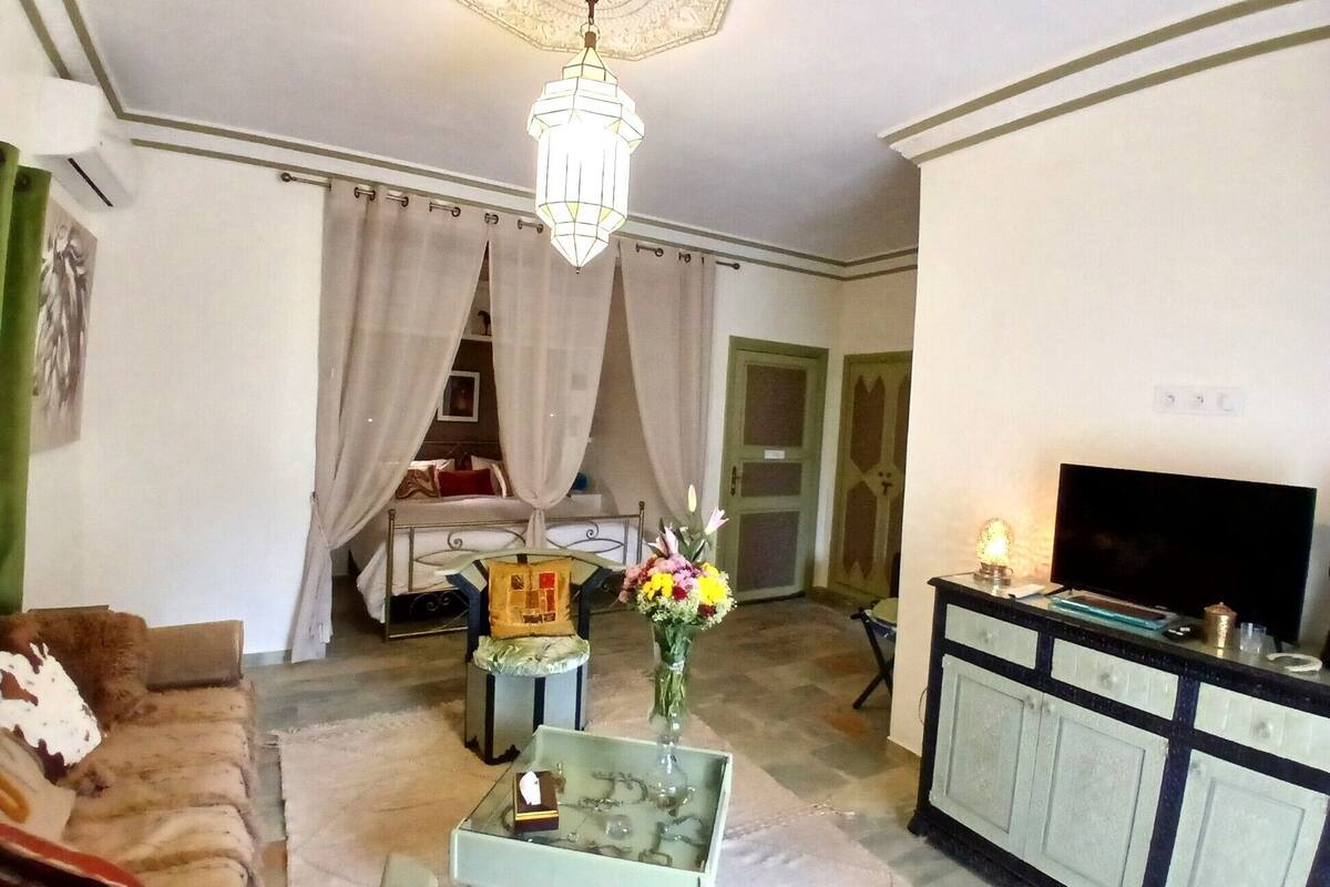 Una sala de estar con sofá, una mesa de centro de vidrio, un mueble para televisión, una araña y una cama con cortinas.