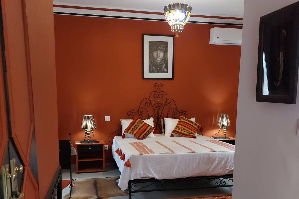 Un dormitorio con una cama, dos mesitas de noche, un candelabro colgante, un aire acondicionado instalado en la pared y una alfombra estampada.