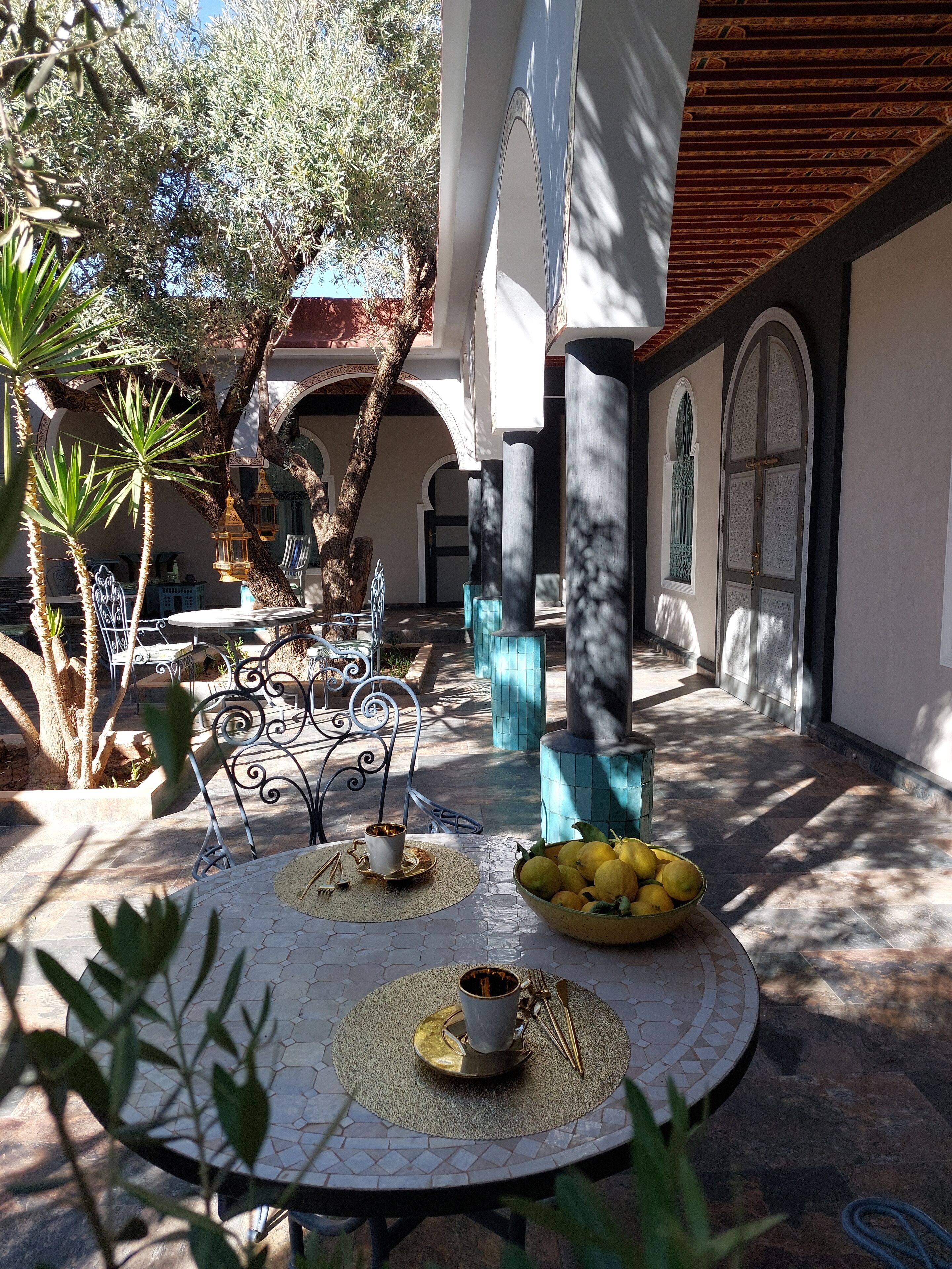 An outdoor patio with a round table set for dining, surrounded by olive trees and a tiled floor.