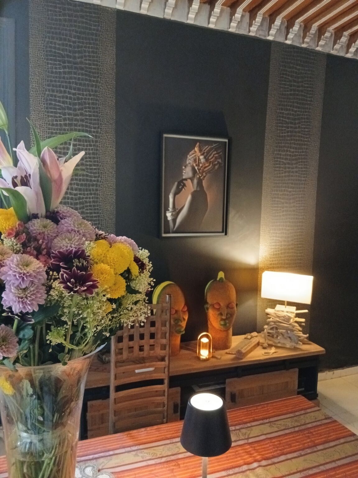 Ein Zimmer mit dunkler Wand, einem gerahmten Kunstwerk, einem Holztisch mit Lampe und einer Blumenkomposition.