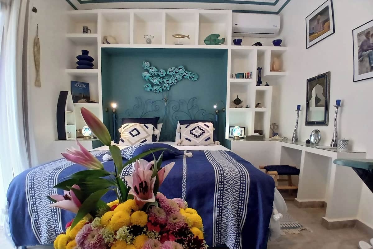Un dormitorio ordenado con cama, mesitas de noche y una estantería en la pared decorada con objetos.