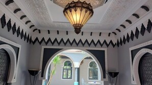 Ein Zimmer mit hoher Decke, aufwendiger Deckenverzierungen und einer großen, dekorativen Leuchte. Zwei Rundbogenöffnungen führen in einen Außenbereich mit Tisch und Stühlen.