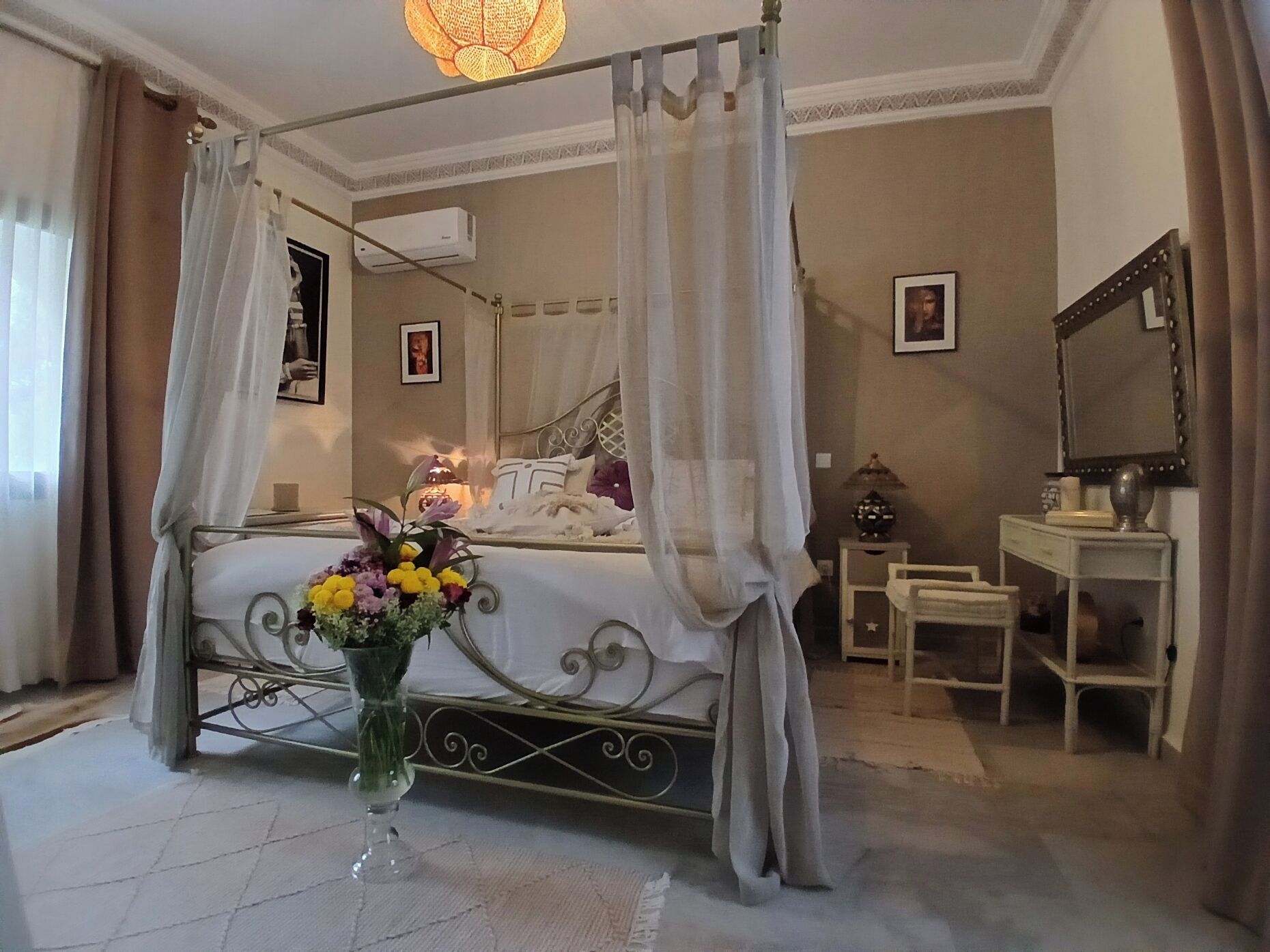 Un dormitorio con cama con dosel, una mesita de noche, un espejo y un jarrón con flores.