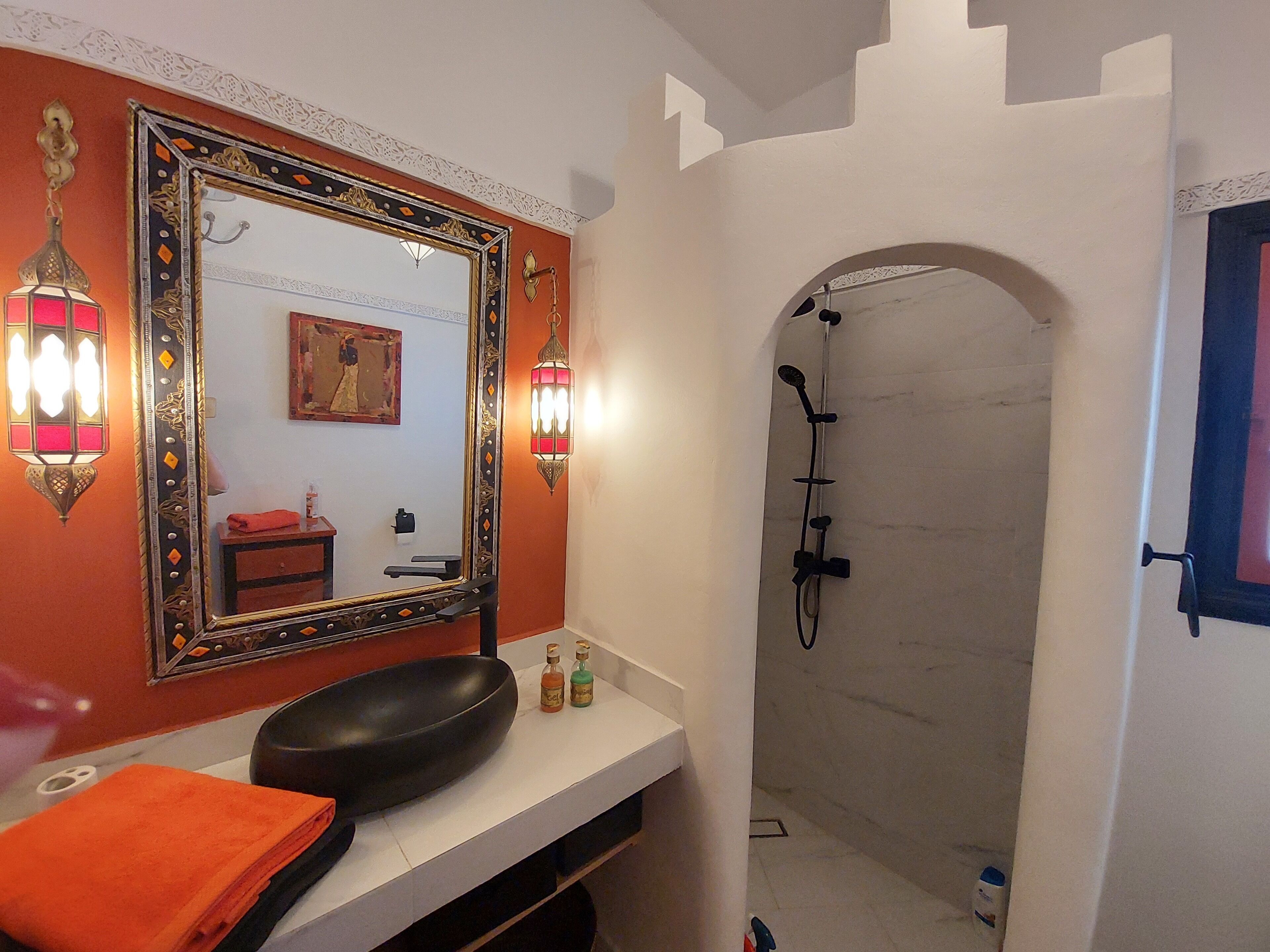 Un baño con un espejo grande y ornamentado, dos luces empotradas en la pared, un lavabo con lavamanos negro y una zona de ducha con arco blanco.